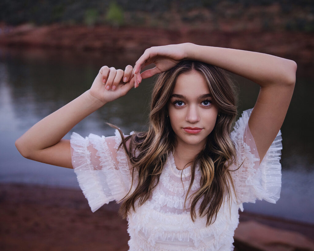 Senior portrait of a teenage girl in a white ruffled dress with arms raised, standing near a mountain lake at dusk