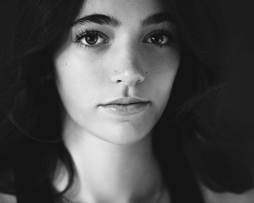 Black and white close-up senior portrait of a teen girl with long lashes and a soft expression, photographed in studio lighting
