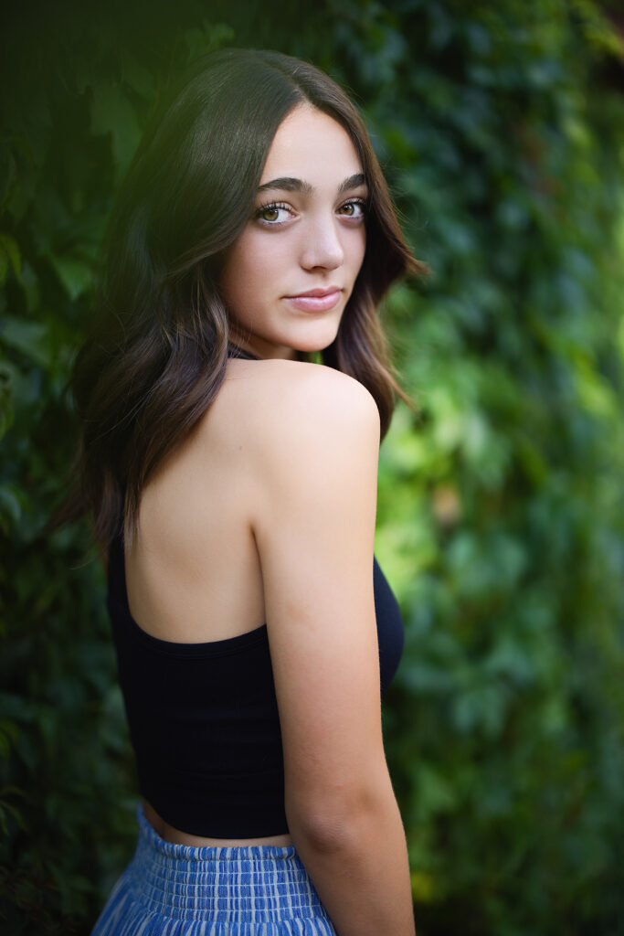 Color outdoor senior portrait of a teen girl in a black top and blue skirt looking over her shoulder against a leafy background