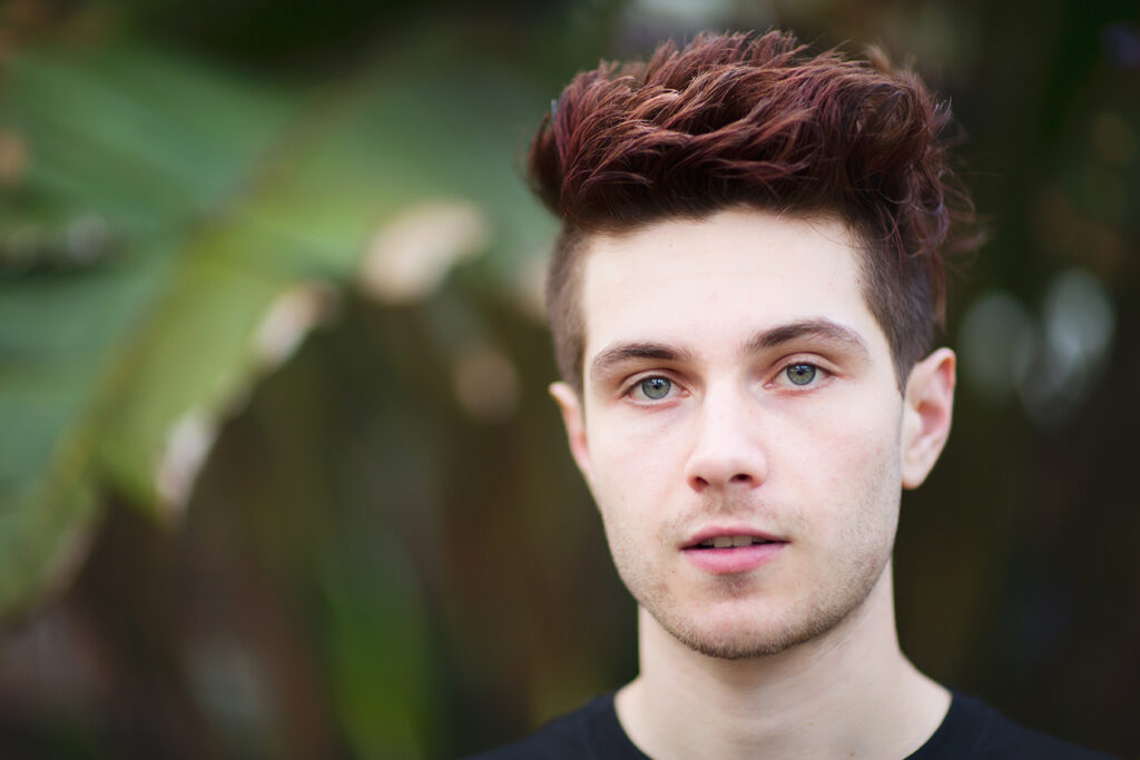 Natural light senior portrait of a teen boy with styled hair and dark shirt, standing in front of tropical greenery