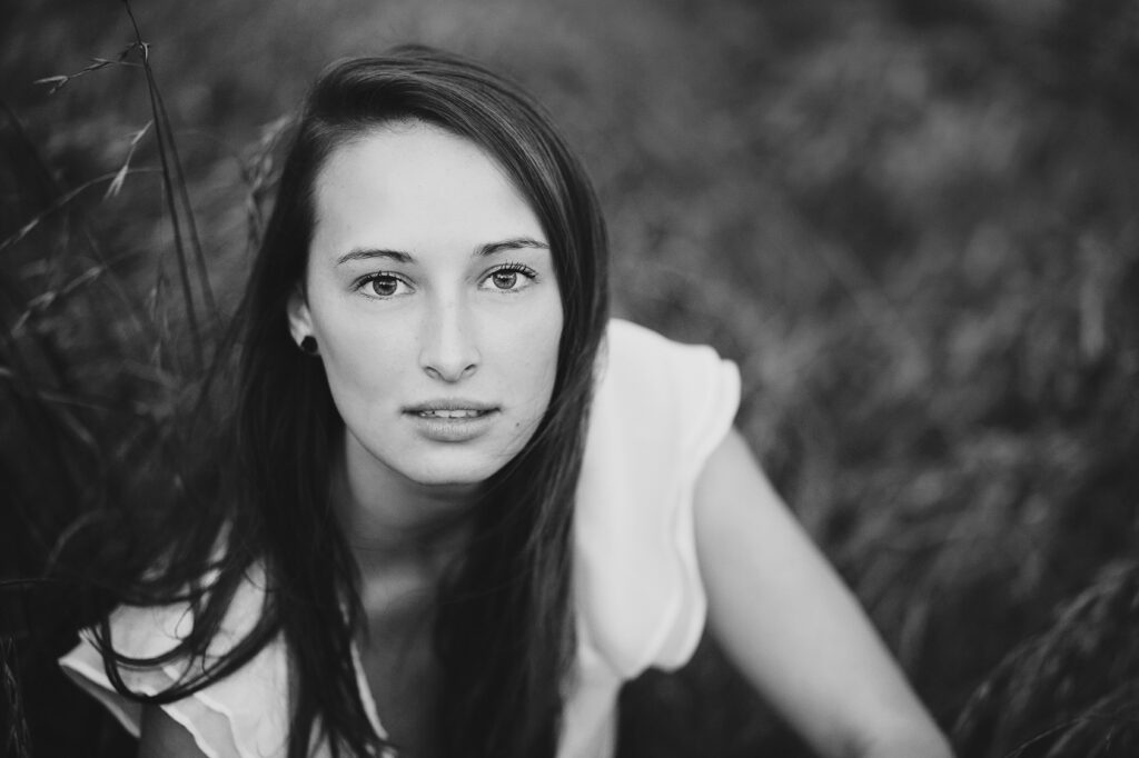 Black and white senior portrait of a teen girl with long dark hair, sitting in tall grass and looking at the camera
