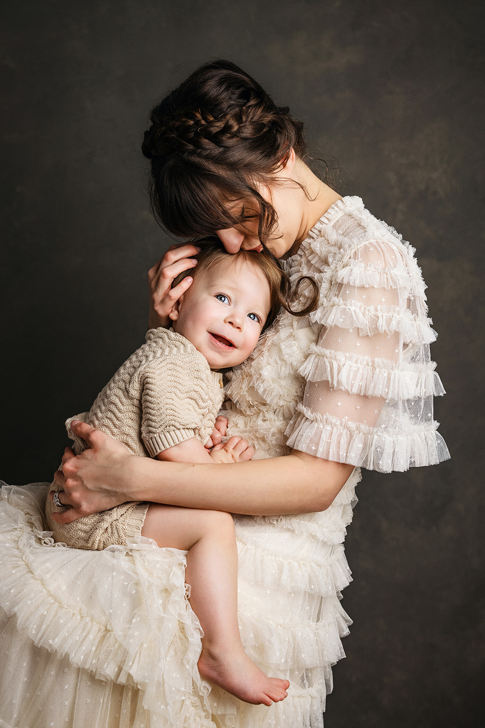 Mother in ruffled ivory dress embraces smiling toddler in timeless motherhood portrait