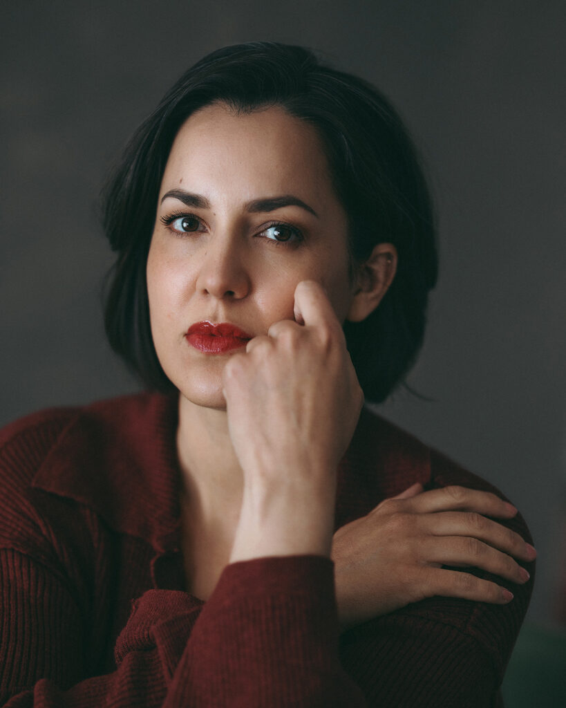 Color portrait of a woman in a red top and red lipstick resting her hand on her face.