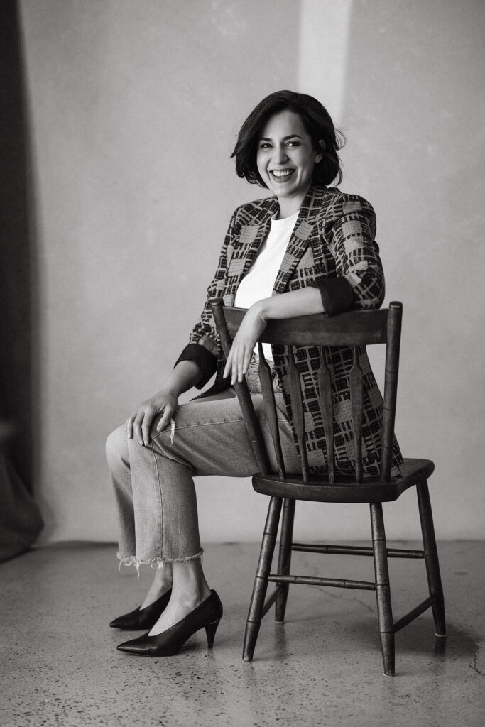 Woman sitting sideways on a wooden chair, smiling warmly in a black and white studio portrait.