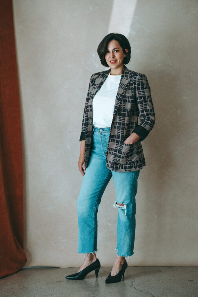 Full-length portrait of a woman in a white tee, plaid blazer, and ripped jeans, smiling in a studio setting.