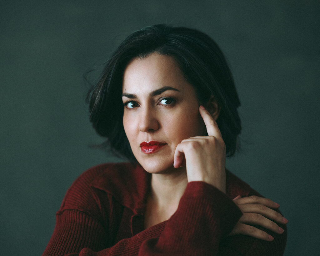 Artistic headshot of a woman in a red sweater resting a finger gently on her cheek with direct eye contact.