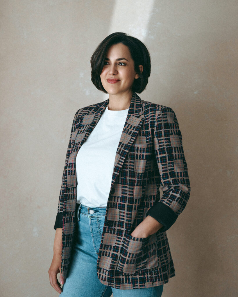 Woman smiling in a patterned blazer and jeans standing against a neutral backdrop.