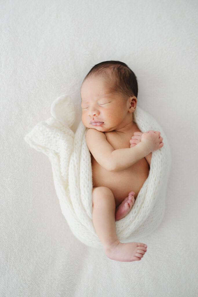 “Newborn baby peacefully sleeping in a cream knit wrap on a white blanket during a Fort Collins newborn photo session.”