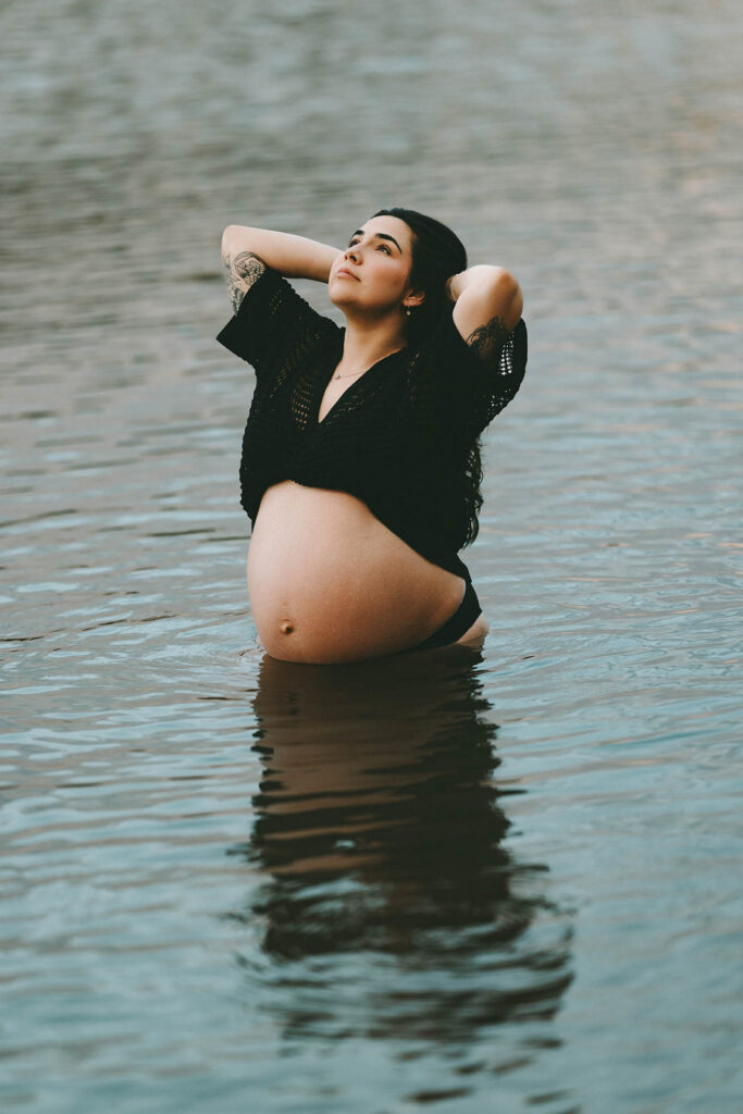 photo of pregnant woman standing in lake and gazing up and pulling her hair back with both hands