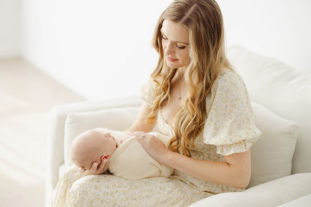 photo of mother sitting in comfortable white chair holding newborn baby in her lap and looking down at him
