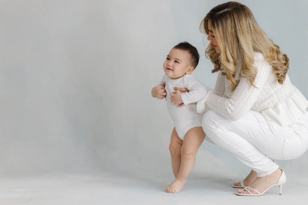 mother helping baby stand and take first steps in studio