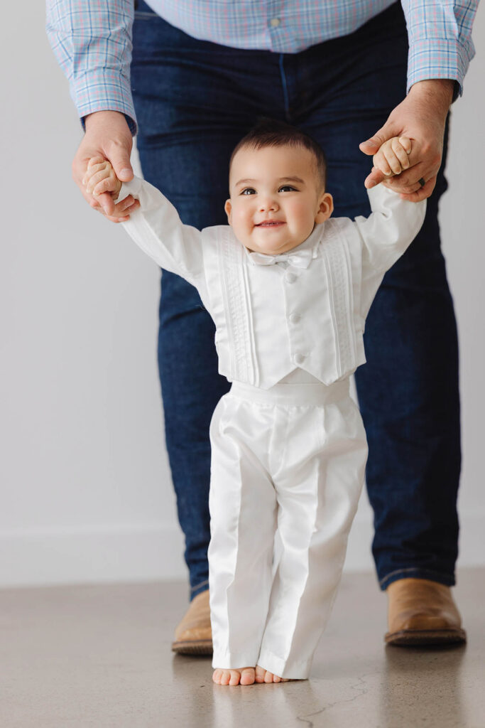 6 month baby standing with support holding parent’s hands during sitter milestone session