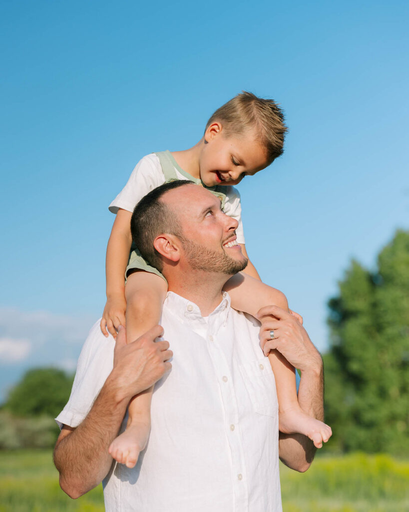 Dad with barefoot boy on his shoulders with blu sky as background, father is looking up at son and son is looking down at dad
