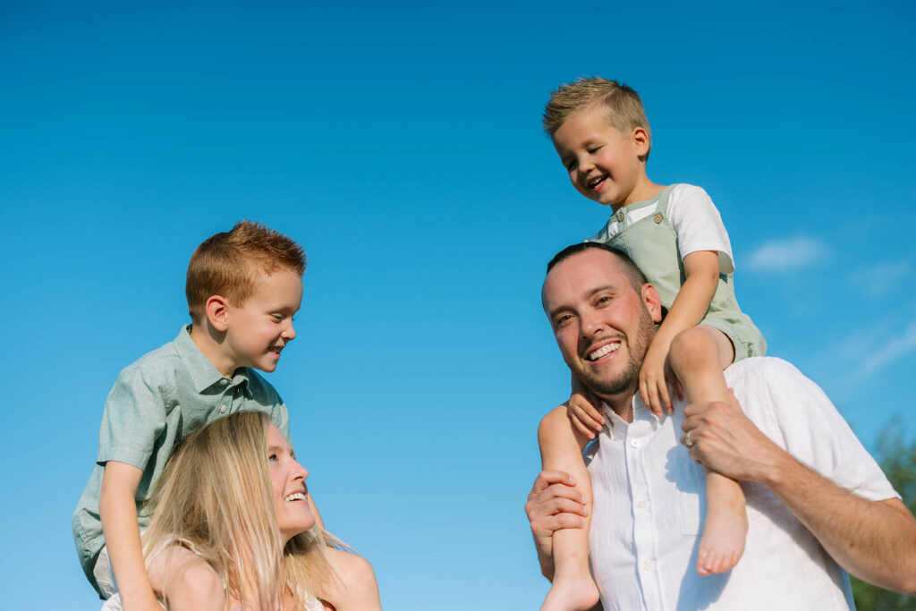 Mom and dad with each with a son on their shoulders with blue sky as background, everyone ids laughing