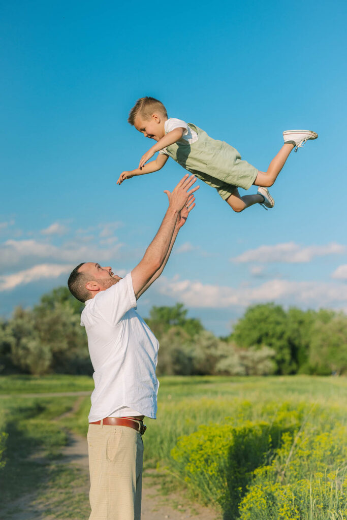 Dad throwing up little boy and catching him with green field and blue skies