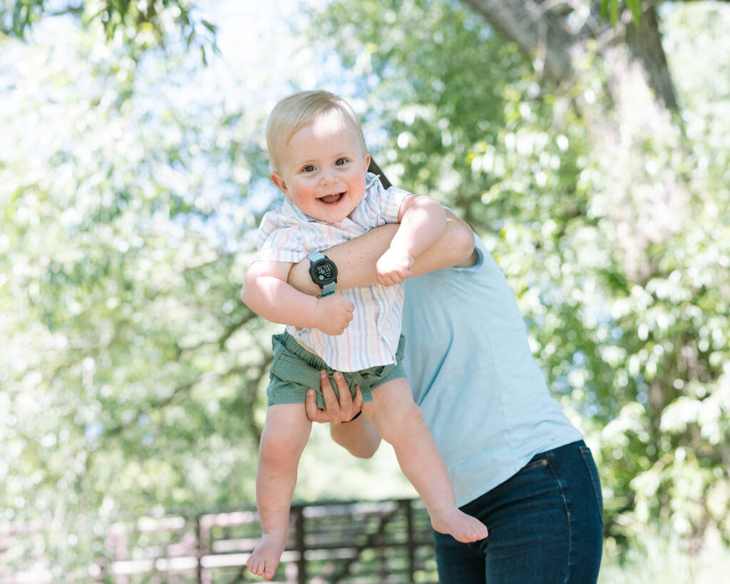 Mother swinging smiling baby toward the camera with a green tree in the background