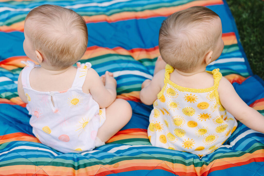 Twin baby girls in sun rompers sitting side by side on a rainbow striped blanket, seen from behind, Fort Collins baby milestone photographer