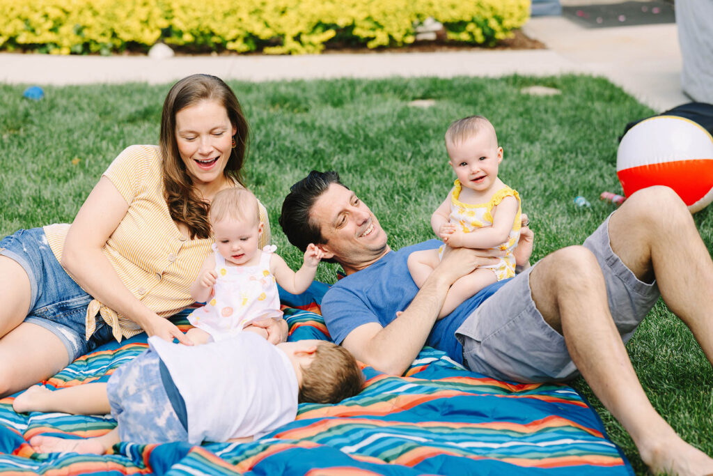 Family of five — mom, dad, toddler boy, and twin baby girls — laughing together on a colorful striped blanket in their backyard during a summer family photography session in Fort Collins, Colorado