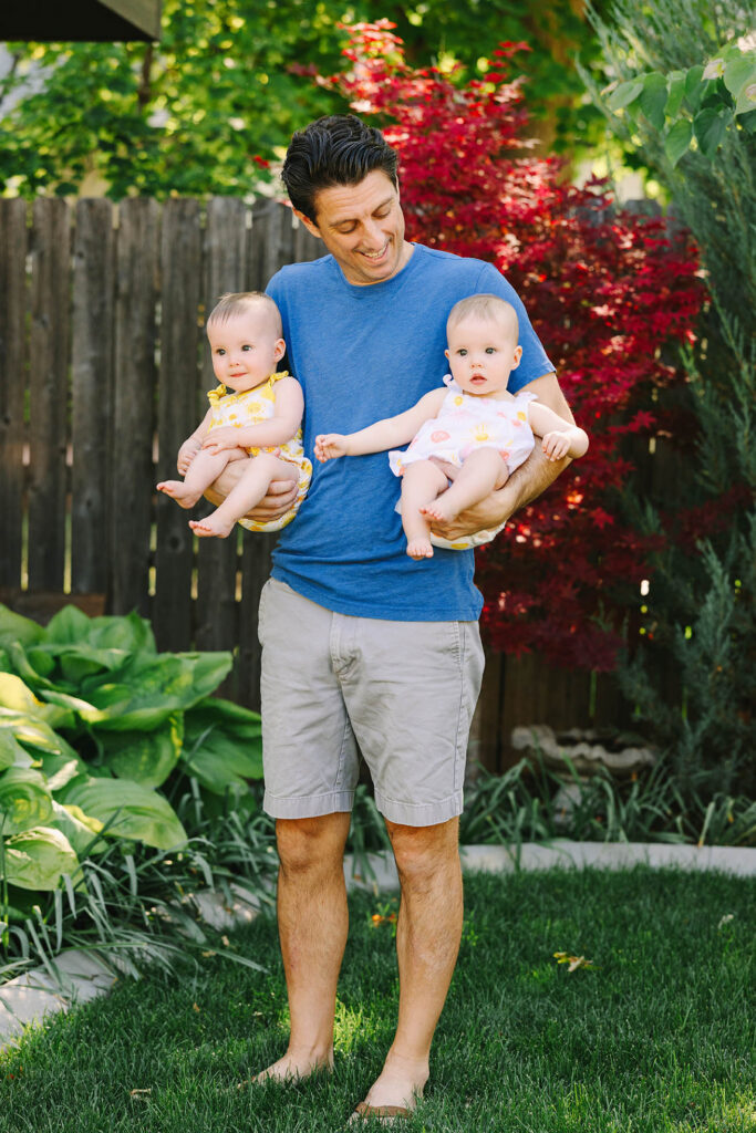 Father holding twin baby girls in matching sunshine rompers in a backyard garden setting, Fort Collins family photographer