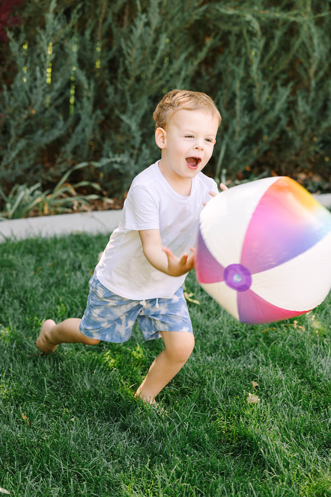 Barefoot toddler boy running across green backyard grass chasing a colorful beach ball, Northern Colorado summer family photos