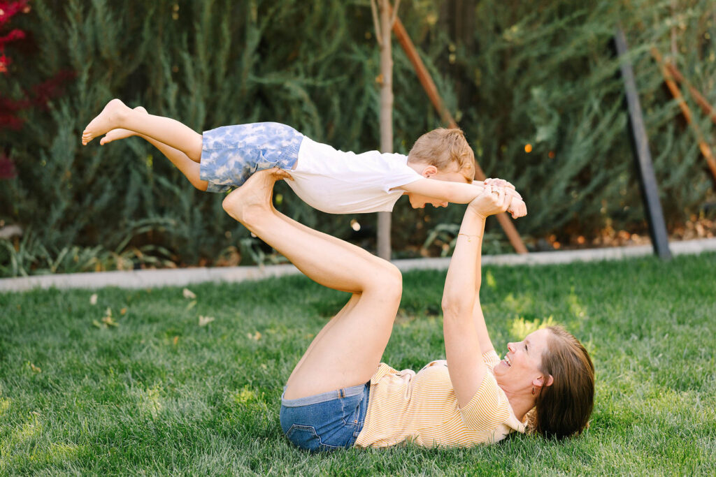 Mother lifting laughing toddler boy overhead while lying on backyard grass, summer family session Fort Collins Colorado