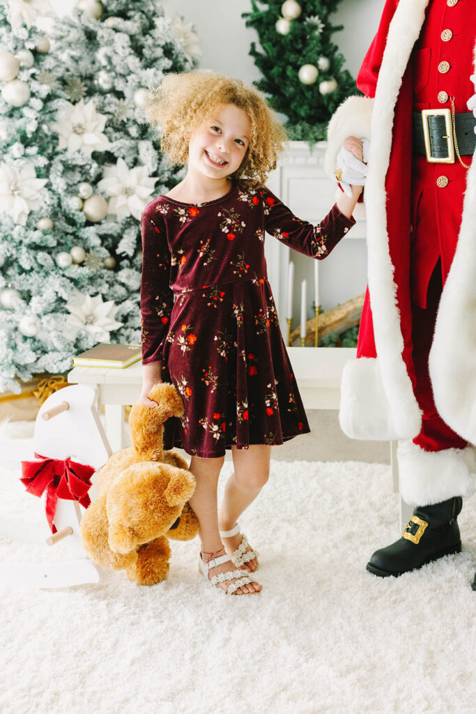 A little girl in a Christmas dress holding Santa's hand and grinning at the camera