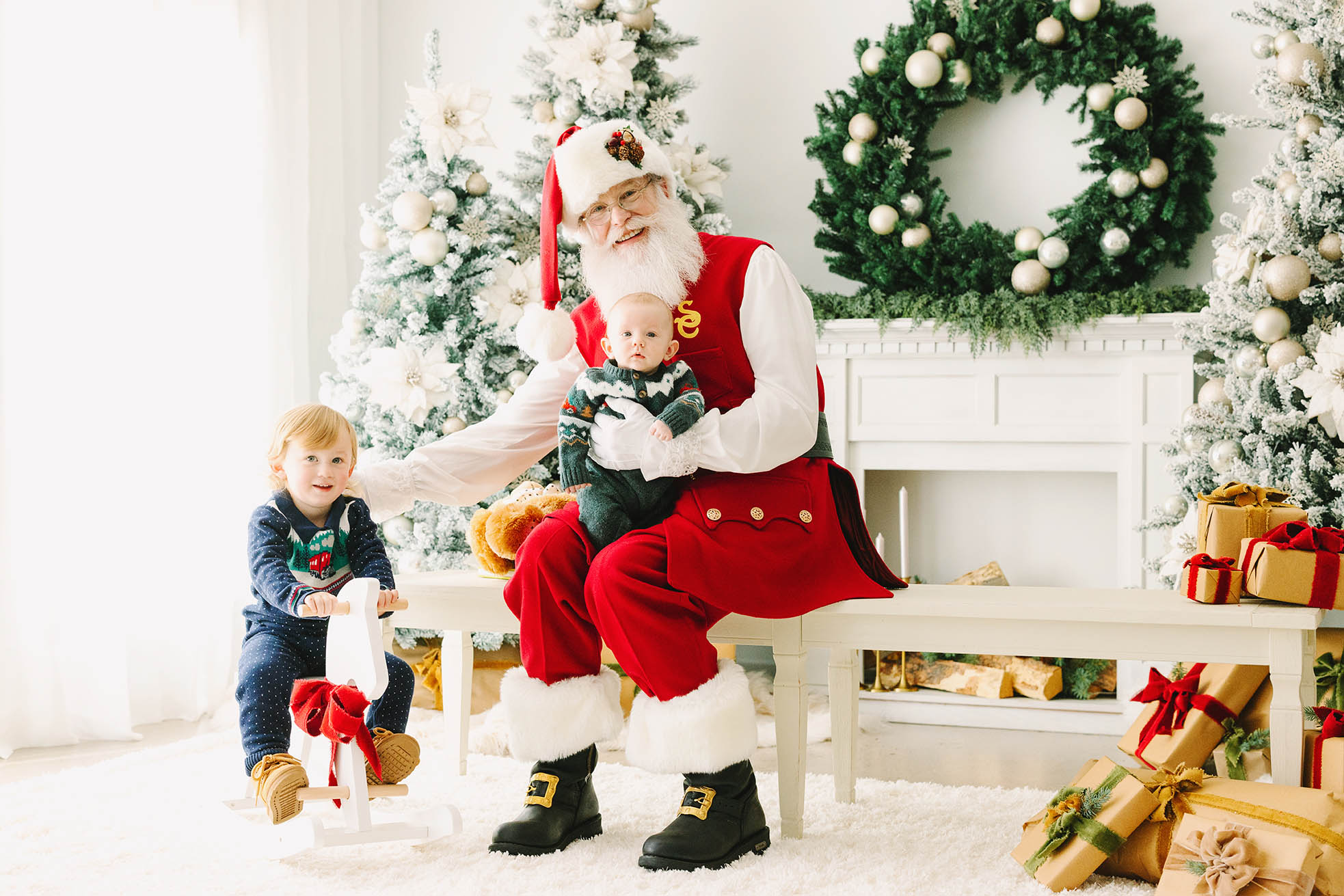 Santa holding a baby and stretching out to put his hand on the back of a boy on a rocking horse