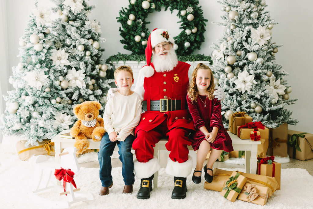 a brother and sister getting their photo taken with Santa at Christmas time in a white Christmas set